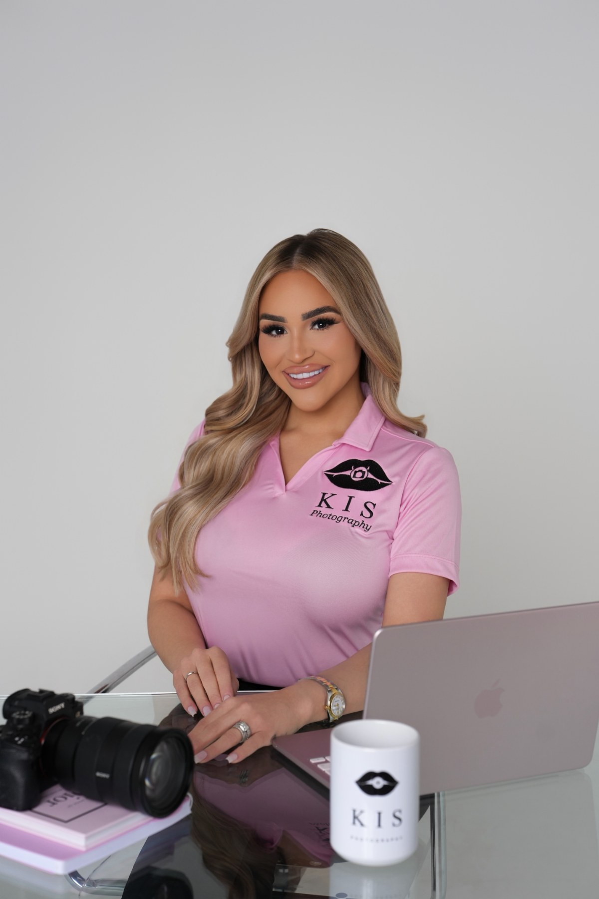 Karina Santucci, owner and photographer of KIS Photography, seated at a desk with a camera and laptop in a pink KIS Photography polo