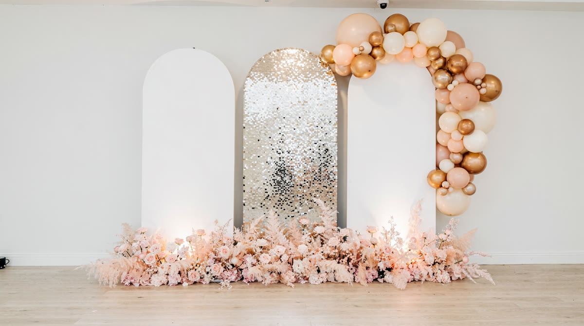 Three arched white backdrop panels with a silver shimmer wall in the center, framed by a blush, cream and gold balloon garland with soft pink florals at the base