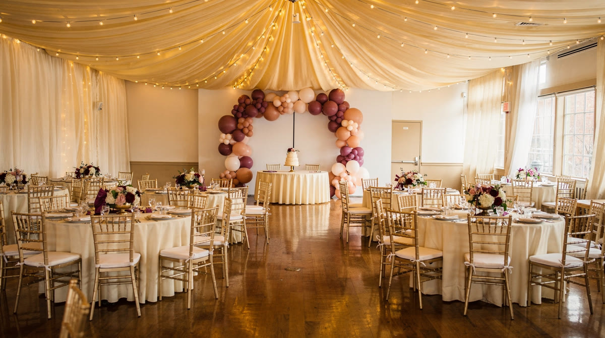 Fully styled event room with ivory ceiling drapes and bistro lights, a mauve and blush balloon garland over the cake table, and round guest tables with gold Chiavari chairs