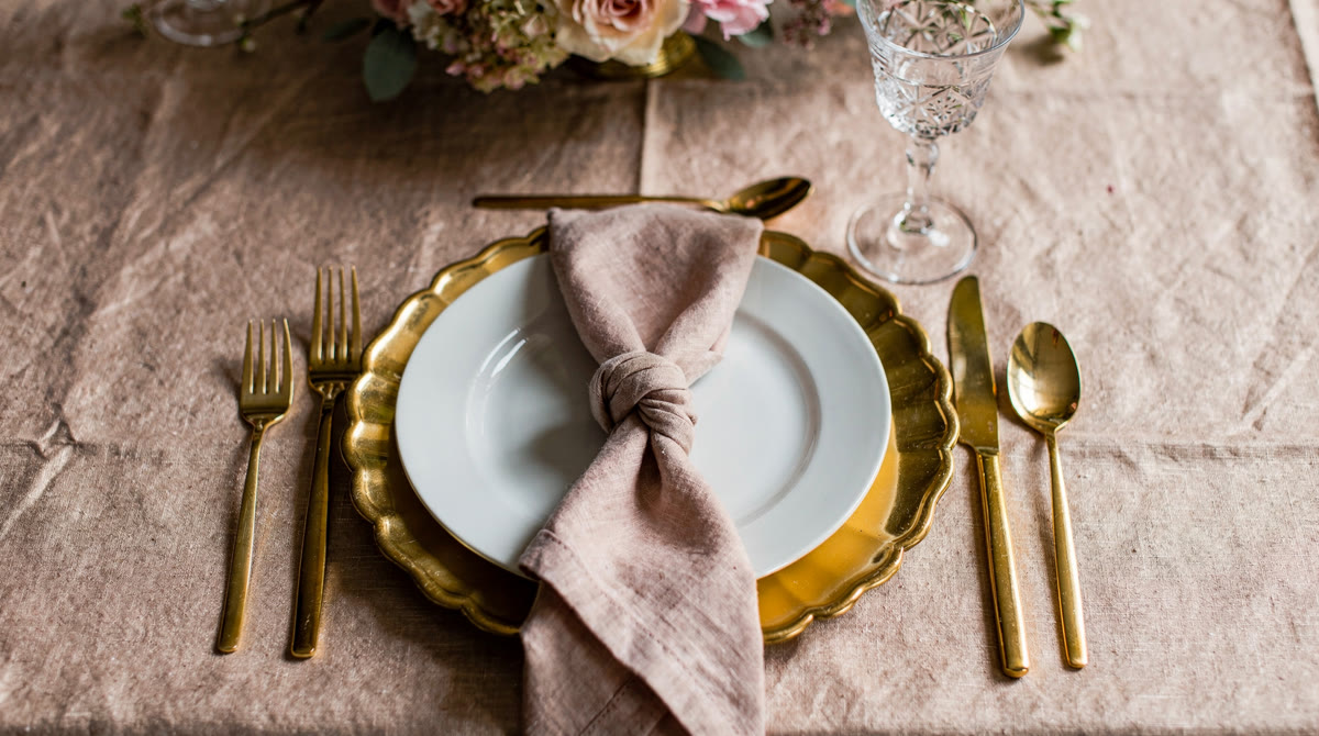 Gold scalloped charger with a white plate, knotted blush linen napkin, gold flatware and a crystal glass on champagne linen