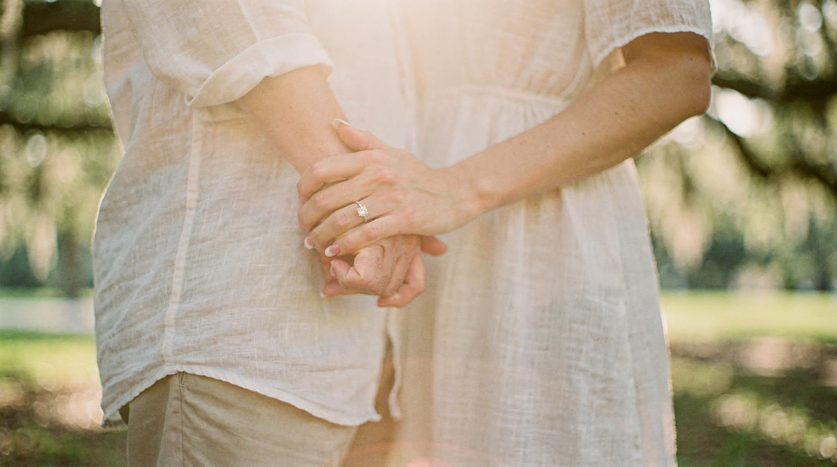 Engaged couple's hands intertwined, engagement ring catching golden-hour light under oak trees