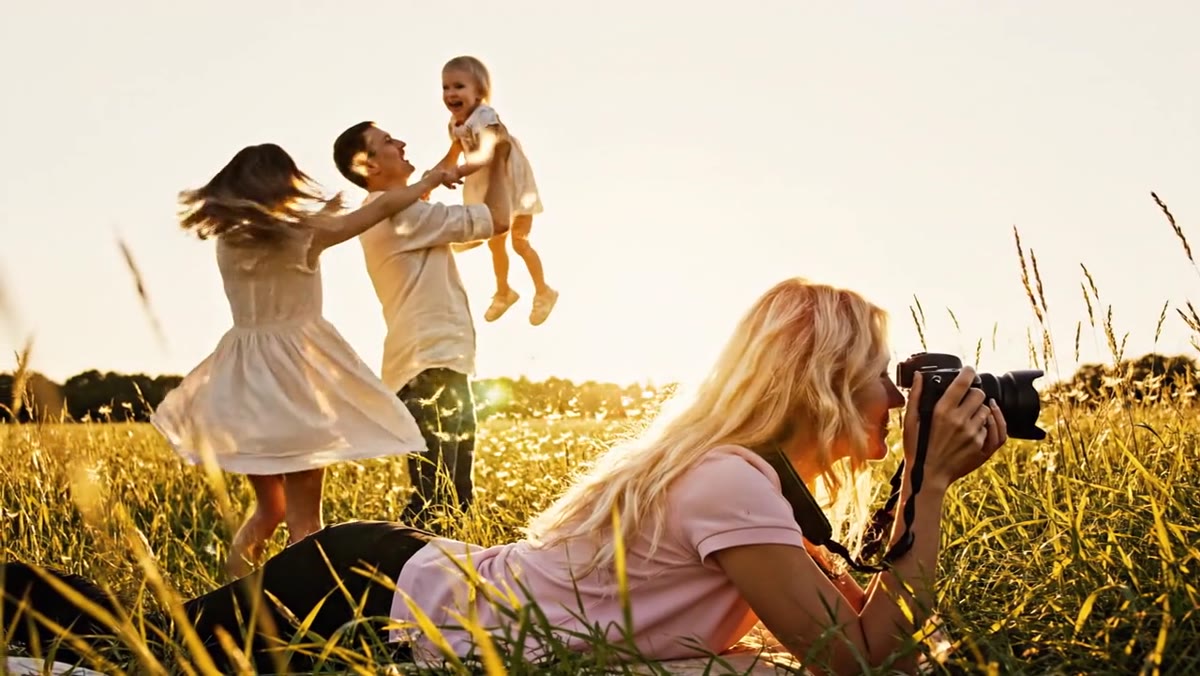 Photographer lying in golden meadow grass photographing a father lifting a toddler while the mother twirls beside them at sunset