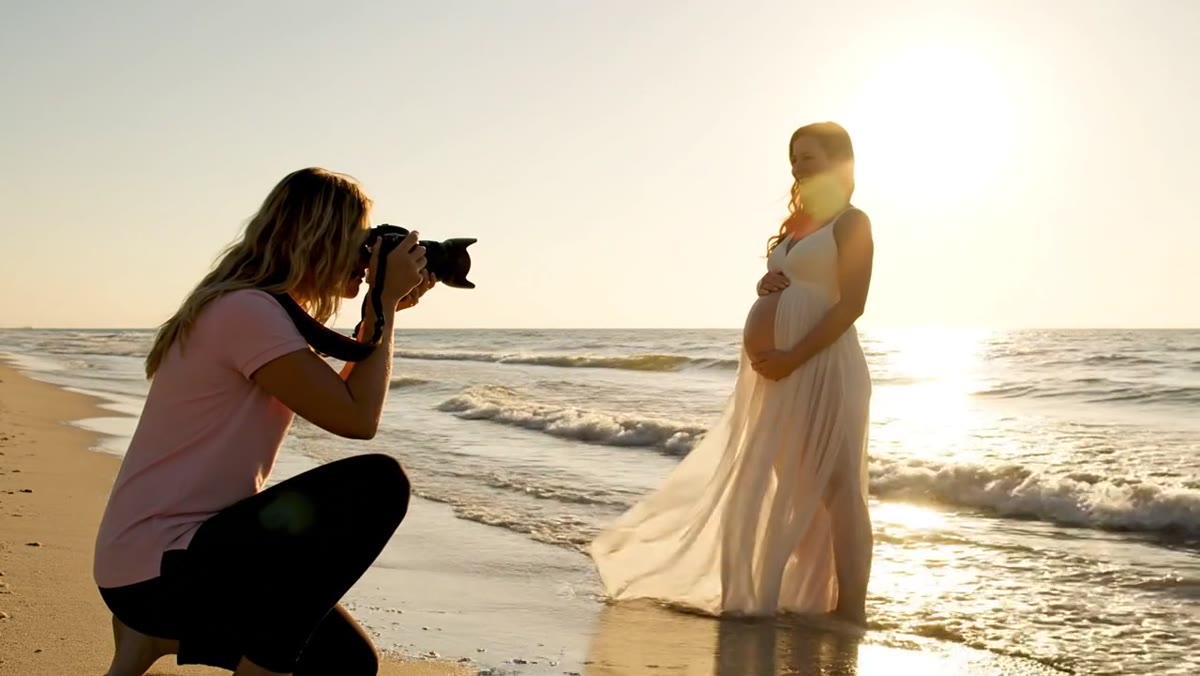 Photographer kneeling in the surf photographing an expectant mother in a flowing cream gown at golden hour